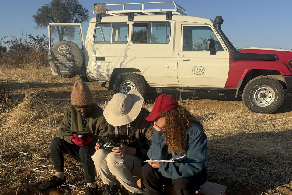 In the foreground three people site on stools, they are looking athe display from a screen attached to a drone remote control as part of a wildlife survey, behind them a long wheel base land rover 4x4 owned by the HALO Trust.