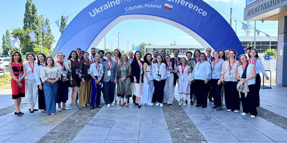 A group of around 30 manly female campaigners stand below an arch reading Ukraine Recovery Conference Gdansk Poland 2026.