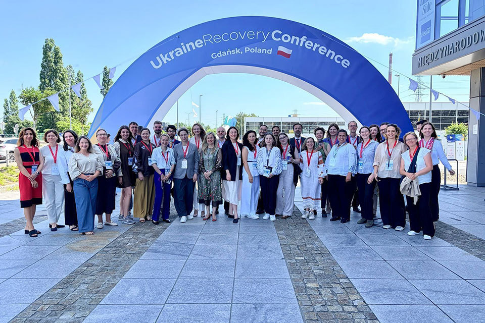 A group of around 30 manly female campaigners stand below an arch reading Ukraine Recovery Conference Gdansk Poland 2026.