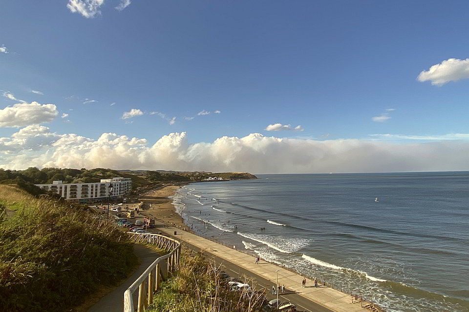 A view looking across a coastal bay, above the horizon, half cliffs and half sea, a cloud of grey smoke can be see drifting from left to right and offshore.