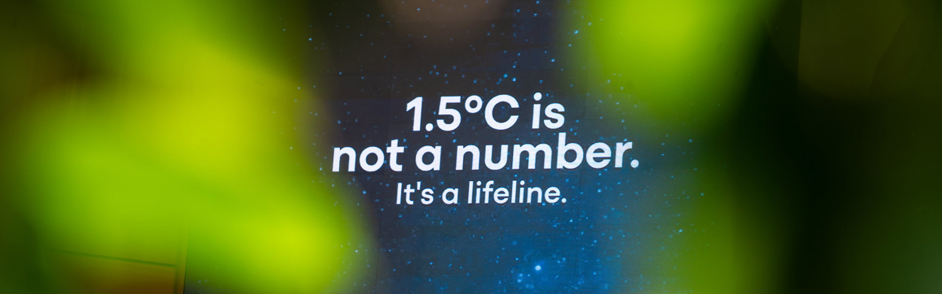 A still from a projection at the UNFCCC intersessional reads "1.5 degrees C is not a number, it's a lifeline. The background comprises bright green blobs, and stars on a navy blue background.
