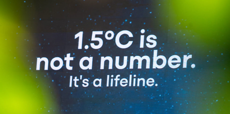 1.5C_960 A still from a projection at the UNFCCC intersessional reads "1.5 degrees C is not a number, it's a lifeline. The background comprises bright green blobs, and stars on a navy blue background.