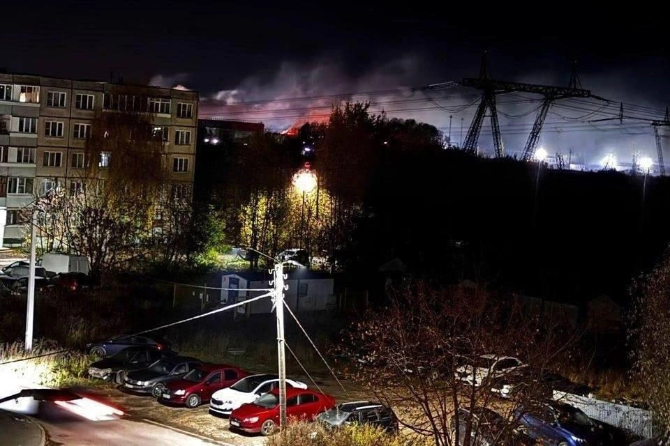 A photo taken at night shows smoke rising behind blocks of flats, in the foreground is a car park in a residential area.